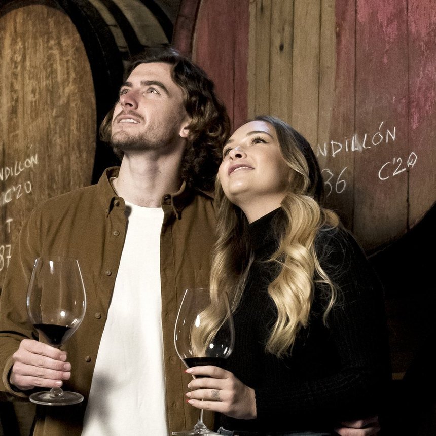 Pareja brindando con copas de vino tinto entre grandes barricas de una bodega